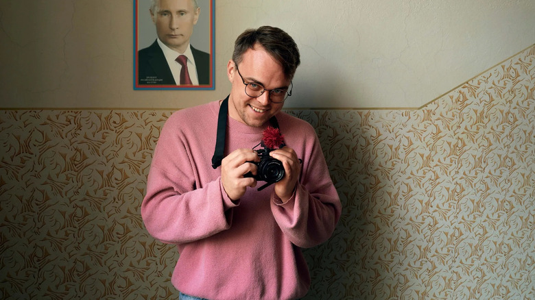 Pavel Talankin affiche un sourire malicieux et tient un appareil photo devant une photo de Vladimir Poutine dans M. Personne contre Poutine