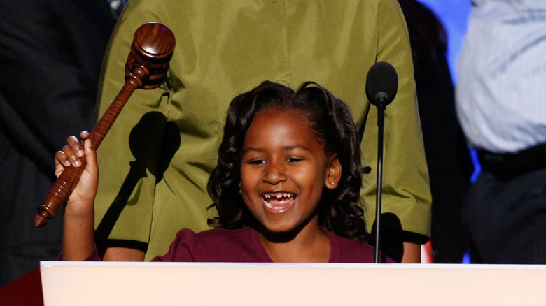 Sasha Obama sourit derrière un micro et tient un marteau
