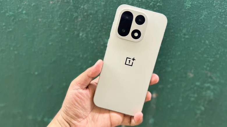 Une main tenant un OnePlus 15 beige devant un mur végétal