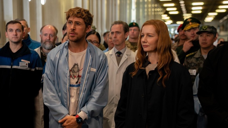 Ryan Gosling dans le rôle de Ryland Grace, debout en tenue décontractée à côté de Sandra Hüller dans le rôle d'Eva Stratt lors d'une réunion bondée du projet Hail Mary