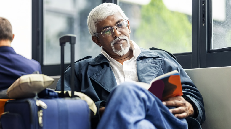 Homme aux cheveux blancs et à la barbe lisant un livre assis en attente dans un aéroport