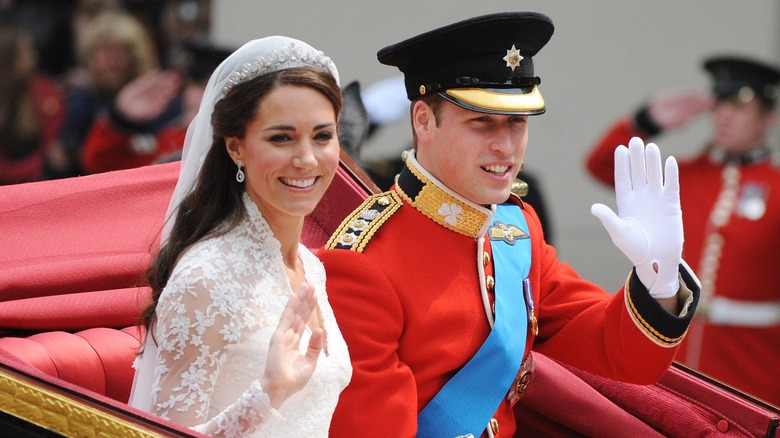 Kate Middleton est assise dans une calèche aux côtés du prince William. Elle porte une robe de mariée blanche en dentelle à col haut avec un diadème et un voile. William porte un uniforme rouge de capitaine et une casquette fourragère.