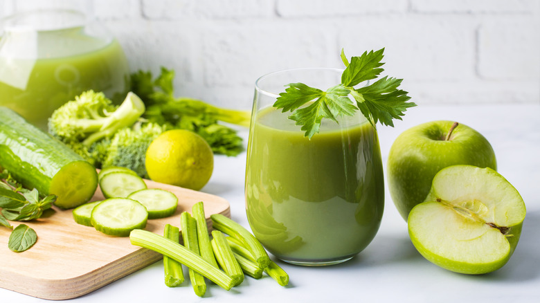 Smoothie vert dans un verre entouré de brocoli, céleri, concombre, citron vert, herbes et pommes vertes