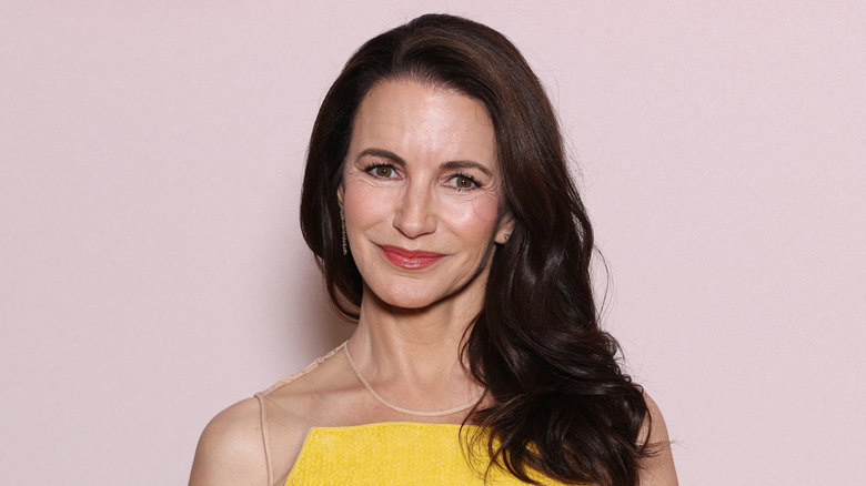Kristin Davis porte une robe jaune vif et une lèvre rouge vif tout en posant devant un fond rose clair