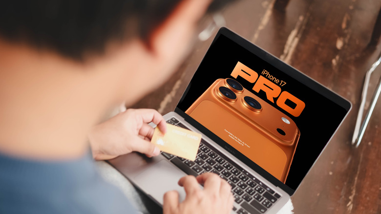 un homme tient une carte de crédit tout en regardant une publicité pour iPhone 17 pro max sur son ordinateur portable