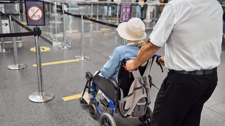 Une femme âgée portant un chapeau est poussée à travers l'aéroport sur un fauteuil roulant par un agent en uniforme.