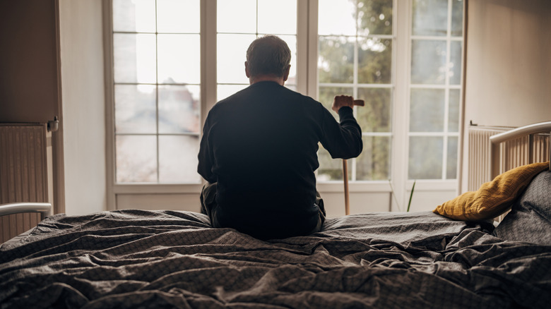 Un homme seul est assis sur un lit face à une fenêtre, tenant une canne à la main