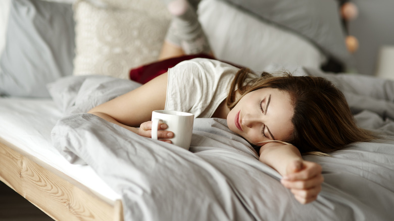 Une personne allongée dans son lit tout en tenant une tasse de café.