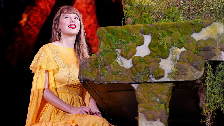 Taylor Swift sourit à la foule en portant une robe jaune et assise devant un piano couvert de mousse.