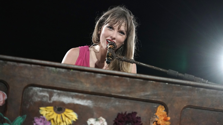 Taylor Swift sourit à la caméra alors qu'elle est assise devant un piano et chante dans un microphone.