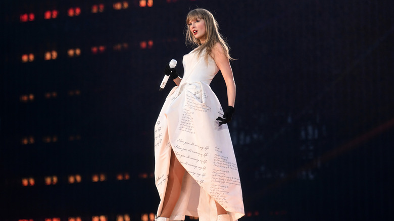 Taylor Swift pose sur scène vêtue d'une robe blanche et de gants noirs, alors qu'elle tient un microphone dans une main.