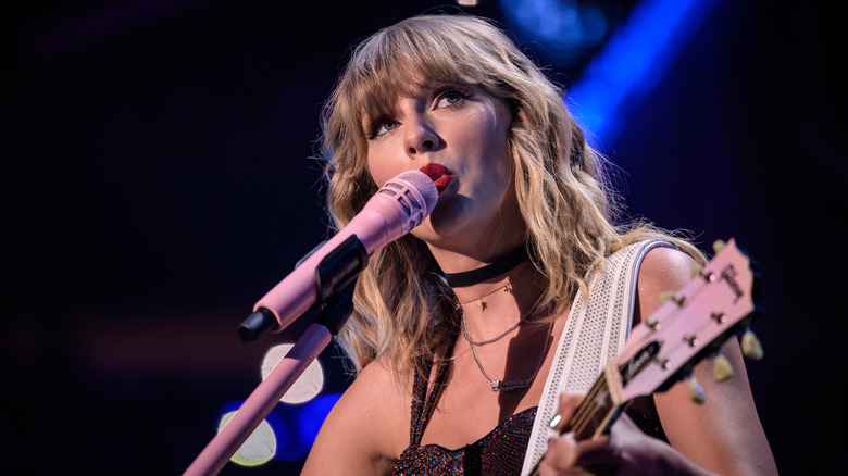 Taylor Swift porte du rouge à lèvres rouge vif alors qu'elle chante dans un microphone rose et joue de la guitare sur scène.
