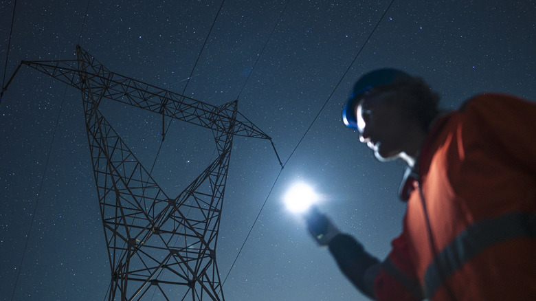 Une personne travaillant dehors la nuit près d’une ligne électrique tout en tenant une lumière vive.