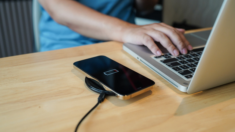Un iPhone plus ancien sur un chargeur sans fil, une personne utilisant un ordinateur portable à côté
