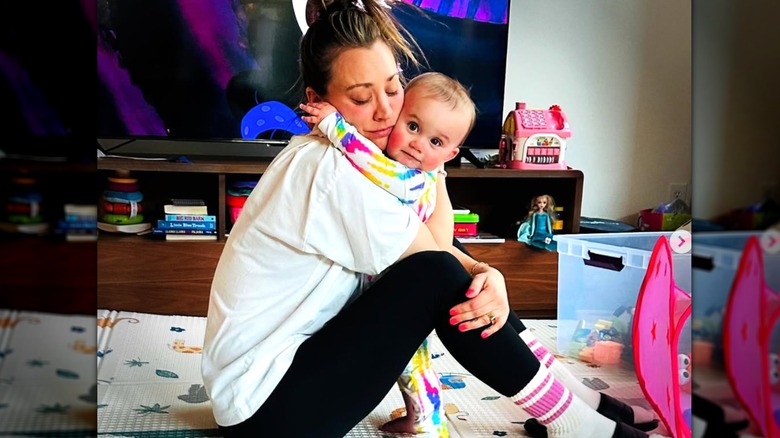 Kaley Cuoco portant des leggings et un T-shirt serrant dans ses bras sa petite fille Matilda, assise sur un tapis avec des jouets autour d'elle.