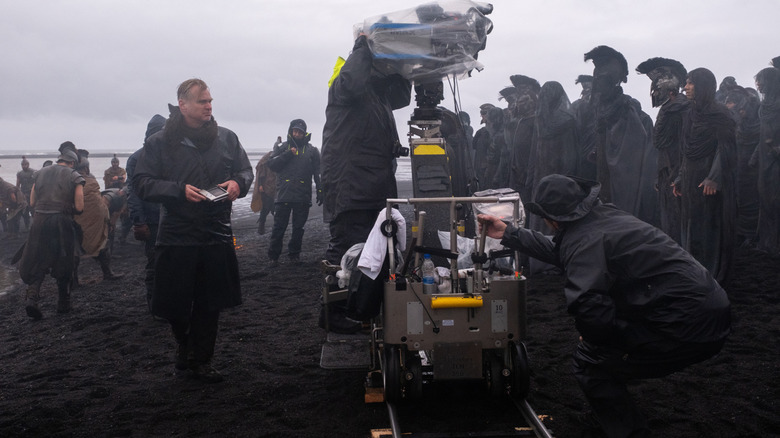 Le réalisateur Christopher Nolan porte une veste de pluie et se tient à côté d'une caméra sur une plage devant plusieurs figurants sur le tournage de L'Odyssée