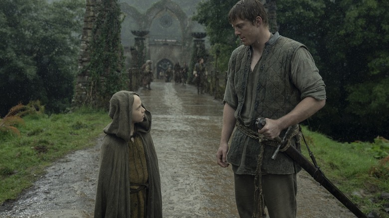 Dexter Sol Ansell dans le rôle d'Egg portant une capuche et debout sur une route sous la pluie à côté de Peter Claffey dans le rôle de Ser Duncan le Grand dans Un Chevalier des Sept Royaumes