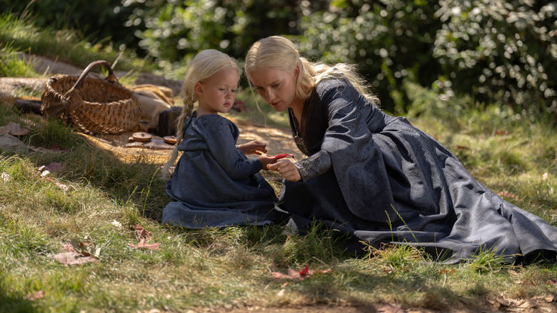Phia Saban dans le rôle d'Helaena Targaryen assise sur une colline herbeuse à côté de Ziggy Barker dans le rôle de Jaehaera Targaryen profitant d'un pique-nique dans la Maison du Dragon