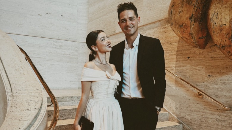 Sarah Hyland dans une robe couleur crème et une queue de cheval lissée dans le dos, debout à côté de Wells Adams dans un costume noir