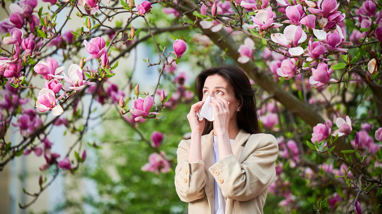 Une femme aux cheveux foncés dans une veste beige se mouche alors qu'elle se tient sous un arbre aux fleurs rosâtres