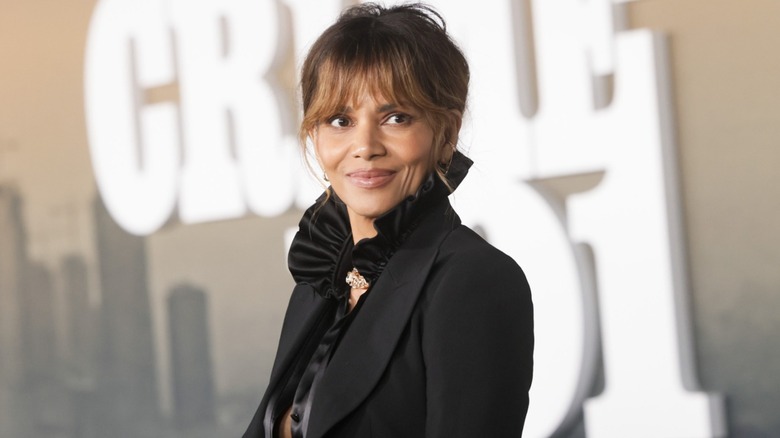 Halle Berry lors d'un événement, les cheveux relevés, vêtue d'une veste noire et d'un chemisier noir à décolleté volanté.