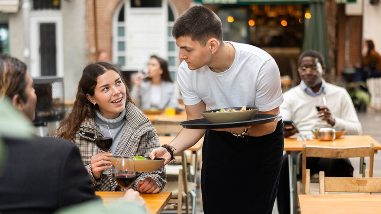 Un serveur apporte des assiettes de nourriture à une cliente assise à l’extérieur d’un restaurant.
