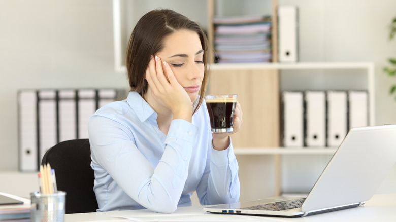 Femme aux yeux fermés devant un ordinateur portable tout en tenant une tasse de café