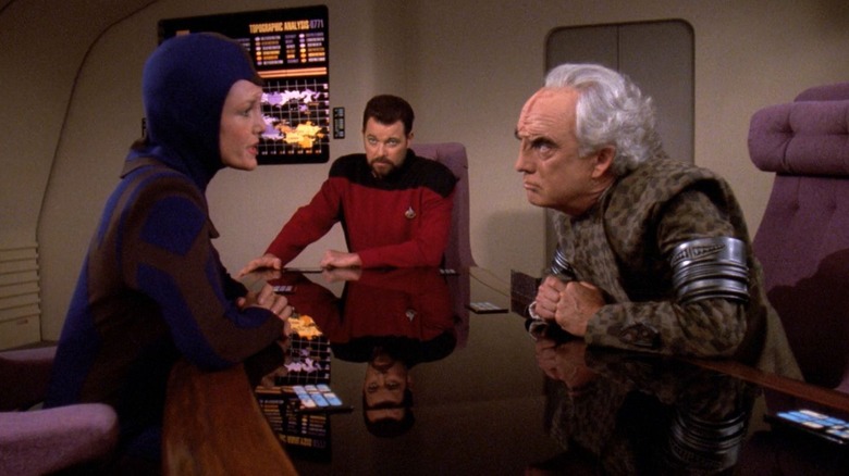 Lorin et Mauric se regardent au-dessus d'une table pendant que Riker regarde Star Trek : La Nouvelle Génération