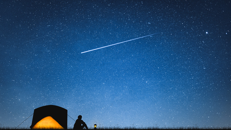 Un campeur assis à côté d’une tente regardant une étoile filante dans le ciel bleu foncé.