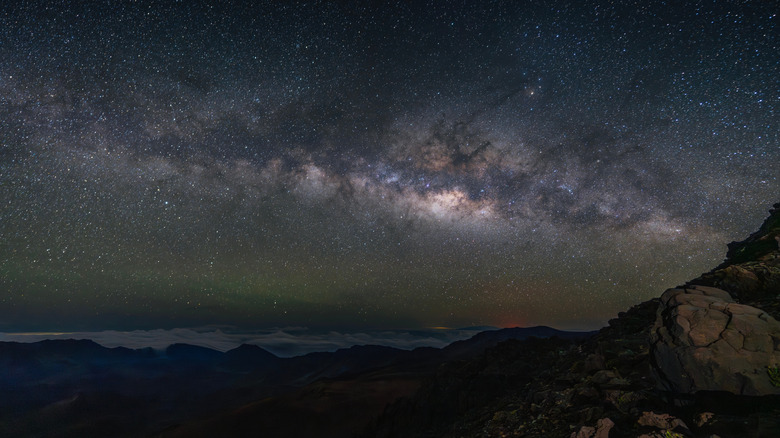 La Voie lactée s'étend brillamment dans le ciel nocturne au-dessus de Haleakalā, illuminant les falaises volcaniques escarpées et une faible mer de nuages ​​en contrebas.