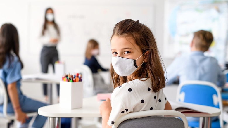 Jeune fille assise en classe avec un couvre-bouche, enseignante, également masquée, en arrière-plan