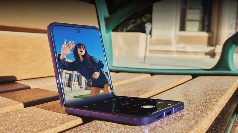 Le Motorola Razr Ultra 2026, à moitié plié sur un banc en bois avec une personne saluant la caméra sur l'écran