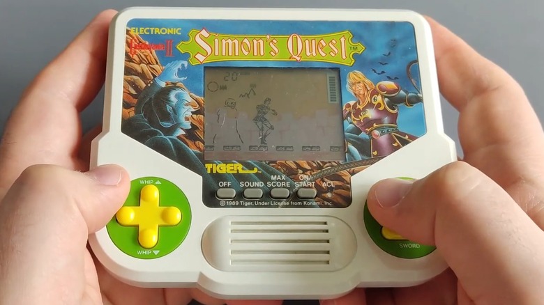 Deux mains tenant et jouant au jeu Tiger Electronics Castlevania II : Simon's Quest sur fond gris