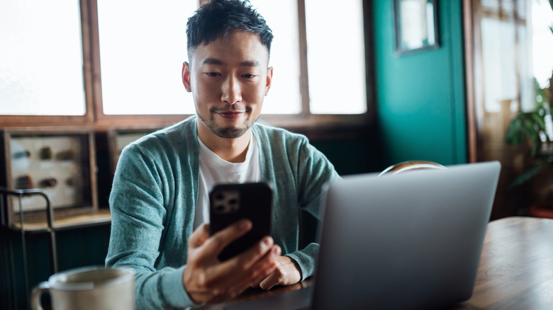 Une personne tenant un téléphone tout en utilisant un ordinateur portable et assise sur un bureau en bois