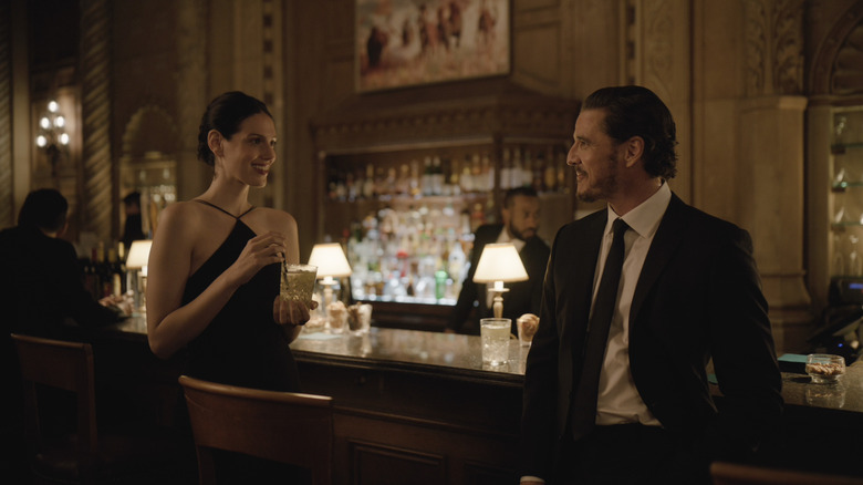 Eva Victor dans le rôle de Nadia Hynes tenant un verre près d'un bar et debout à côté de Pedro Pascal dans le rôle d'Alex Serian dans Behemoth !