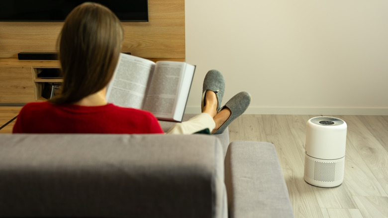 Une femme en chemise rouge est assise sur un canapé en train de lire un livre tandis qu'un purificateur d'air est posé par terre à côté d'elle