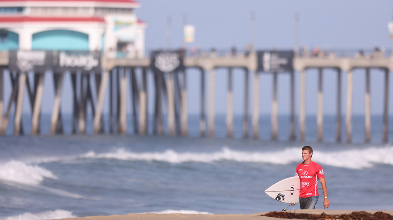 Surfeur en rouge portant une planche avec des vagues floues et une jetée en arrière-plan