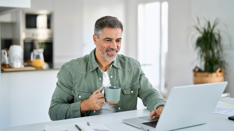 homme aux cheveux gris sur un ordinateur portable en train de prendre un café