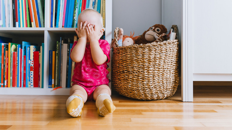 Une petite fille est assise devant une étagère colorée à côté d'un panier de jouets couvrant son visage